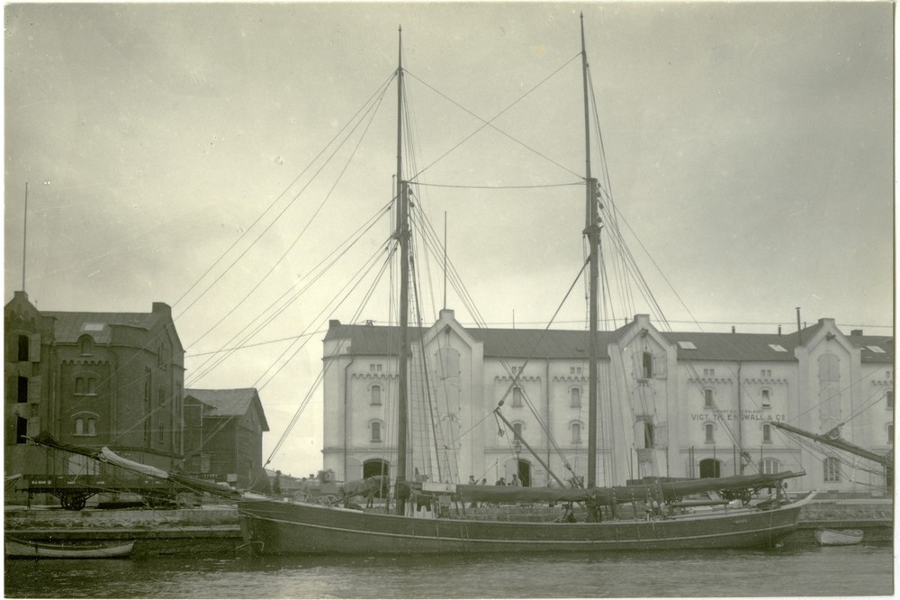 Det tvåmastade segelfartyget Agnes ligger förtöjd vid en kaj. I bakgrunden på det svartvita fotografiet syns äldre magasinsbyggnader.