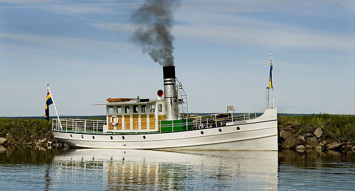 Flottisten är en vitmålad bogserbåt med en styrhytt och en skorsten med svart rök midskepps. Överbyggnaden har sju synliga fönsterrutor. På bilden ligger flottisten vid strandkanten på en sjö. 