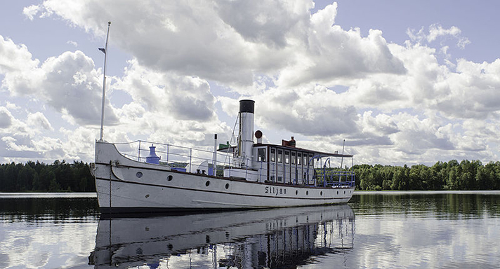 Siljan är en vitmålad varpbåt med skorsten midskepps och styrhytt akter om den. På bilden ligger fartyget i en stilla insjö.