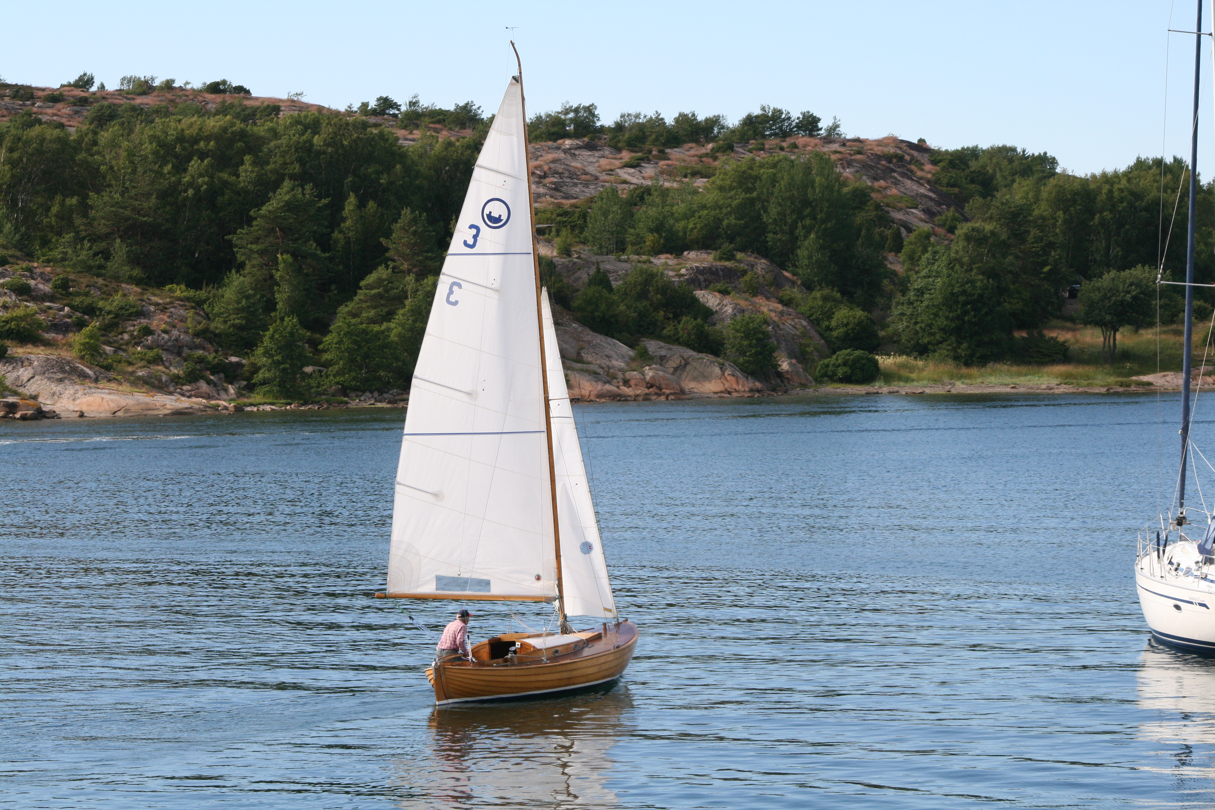 Anzy är en segelbåt med satta vita segel. Skrovet är av ek och klinkbyggt och en man sitter i sittbrunnen och styr båten bort från kameran. 