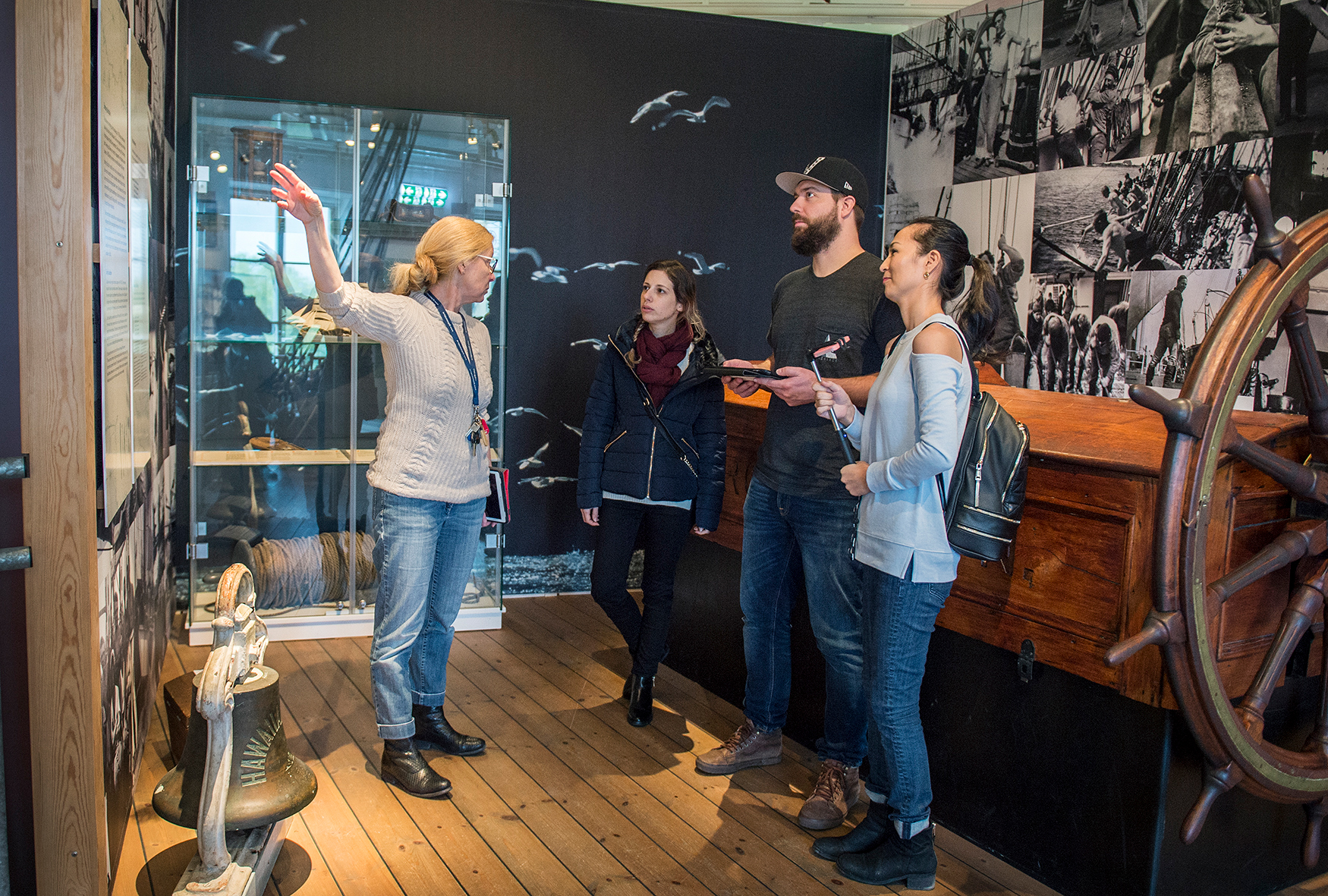 Pedagog berättar för vuxna SFI- elever om livet ombord  segelfartyg. De står i en utställning av ett segelfartyg.